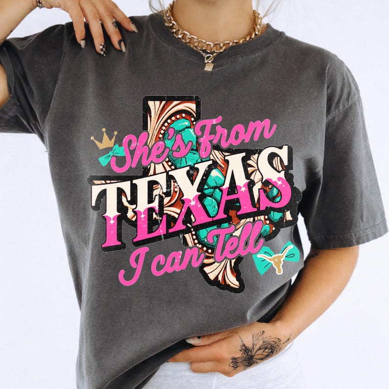 Peut inclure: T-shirt gris fonc&eacute; avec l'inscription "She's From TEXAS I Can Tell" en rose et blanc, et un motif en forme du Texas turquoise et marron. Le t-shirt a un aspect vintage et us&eacute;.
