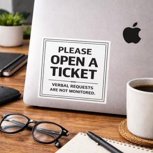 May include: A white sticker on a laptop with the text "PLEASE OPEN A TICKET. VERBAL REQUESTS ARE NOT MONITORED." The laptop has a black Apple logo. The sticker is on a wooden desk with a cup of coffee, glasses, and a notepad.