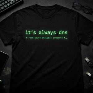 May include: Black t-shirt with the text "it's always dns" in a green, computer-like font. The phrase "# root cause analysis complete #_" is below the main text. The shirt is on a dark surface with tech accessories.