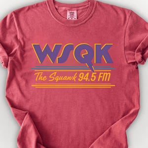 May include: A maroon t-shirt with the text "WSQK The Squank 94.5 FM" in purple, yellow, and blue. The shirt has a crew neck and short sleeves, and the fabric is slightly wrinkled.