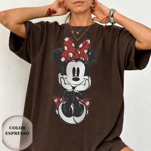 Puede incluir: Camiseta color espresso con un gr&aacute;fico desgastado de Minnie Mouse. Minnie est&aacute; representada en blanco y negro con un lazo rojo de lunares. La camiseta tiene un corte holgado y una est&eacute;tica vintage.