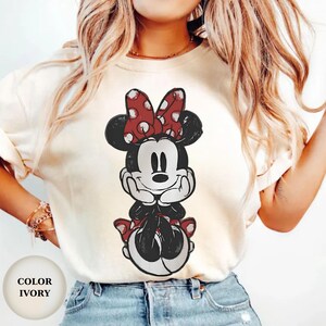 Puede incluir: Camiseta color marfil con un gr&aacute;fico desgastado de Minnie Mouse. Minnie est&aacute; representada en blanco y negro con un lazo rojo de lunares y una falda a juego. El color de la camiseta est&aacute; etiquetado como "IVORY" en un c&iacute;rculo en la parte inferior izquierda.