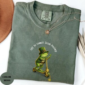 May include: A moss-colored t-shirt with the phrase "Off to Commit Irish Foolery" encircling a cartoon frog riding a scooter. The frog wears a green hat and jacket. A gold-colored necklace with a pendant is draped across the shirt.