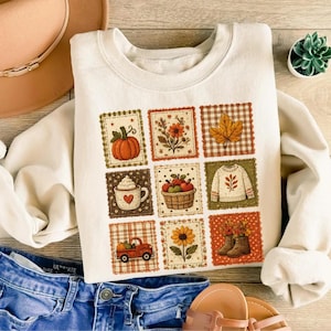 Op de afbeelding: Een crèmekleurig sweatshirt met ronde hals voorzien van een raster van negen vierkante illustraties met herfstthema's, waaronder een pompoen, een mok cacao, een rode vrachtwagen, een trui, een zonnebloem en herfstbladeren in warme tinten.