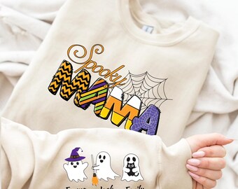 May include: A beige sweatshirt featuring the text "Spooky Mama" in a Halloween design. The word "Mama" is decorated with candy corn and spiderweb patterns. The sleeve displays three cartoon ghosts with the names Emma, Jack, and Emily.