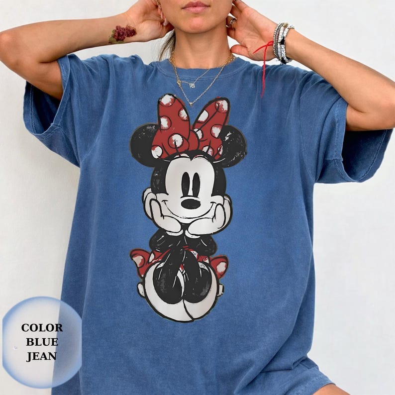 Puede incluir: Camiseta azul vaquero con un gr&aacute;fico desgastado de Minnie Mouse. Minnie Mouse est&aacute; representada con un lazo rojo con lunares blancos. La camiseta tiene las palabras "COLOR BLUE JEAN" impresas.