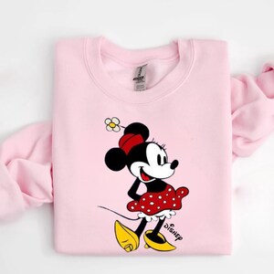 Puede incluir: Sudadera rosa claro con un dise&ntilde;o de Minnie Mouse. Minnie lleva su atuendo cl&aacute;sico: una falda roja de lunares, zapatos amarillos y un lazo rojo. La palabra "Disney" est&aacute; impresa en negro debajo.