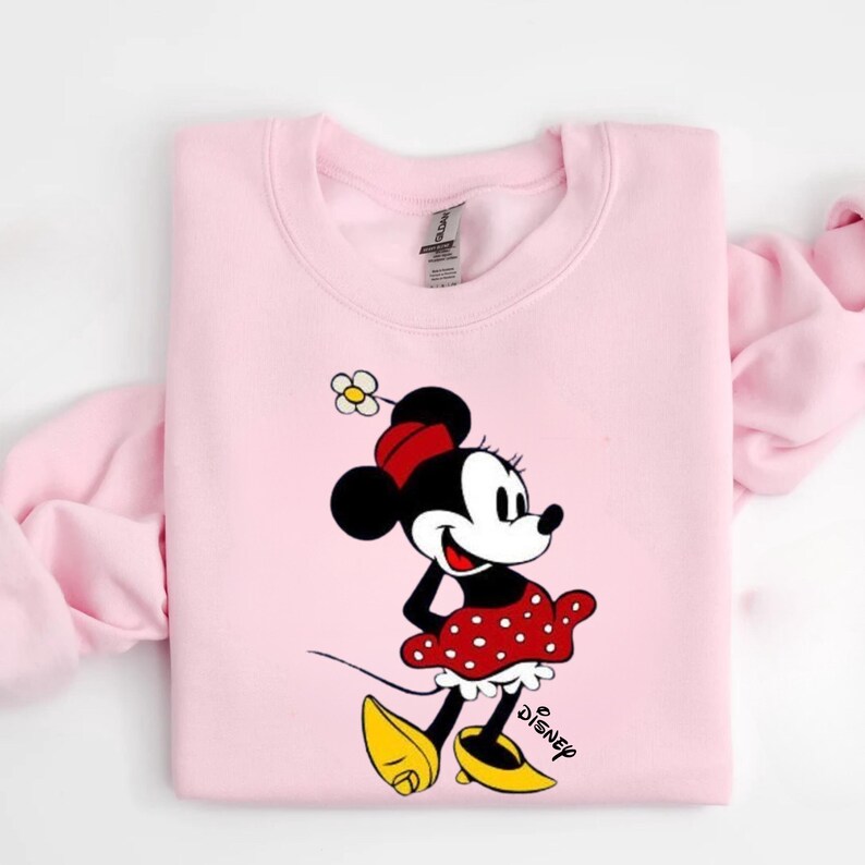 Puede incluir: Sudadera rosa claro con un dise&ntilde;o de Minnie Mouse. Minnie lleva su atuendo cl&aacute;sico: una falda roja de lunares, zapatos amarillos y un lazo rojo. La palabra "Disney" est&aacute; impresa en negro debajo.
