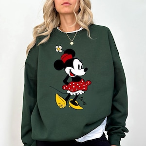 Puede incluir: Sudadera verde oscuro con un gr&aacute;fico de Minnie Mouse. El personaje de dibujos animados lleva una falda roja de lunares, un lazo rojo y zapatos amarillos. La palabra "Disney" est&aacute; impresa debajo.