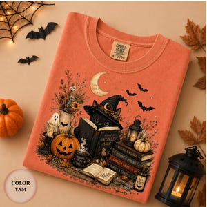 May include: An orange crewneck t-shirt featuring a graphic print of a black cat wearing a witch hat while reading a book titled A Guide to Halloween Magic. The design includes pumpkins, a crescent moon, bats, lanterns, and stacked books.