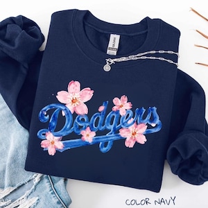 May include: A navy blue crewneck sweatshirt featuring the word Dodgers in a blue watercolor script font, accented with pink cherry blossom illustrations. A silver chain necklace with a circular pendant rests near the neckline of the garment.