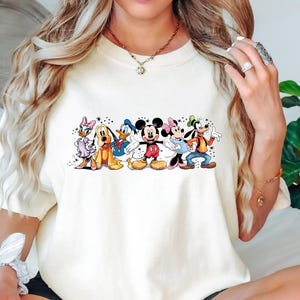 Puede incluir: Camiseta de color marfil con un gr&aacute;fico colorido de personajes de Disney, incluyendo Mickey Mouse, Minnie Mouse, Donald Duck, Daisy Duck, Goofy y Pluto. La camiseta tiene las palabras "Super Soft" y "Ships Next Day" impresas.