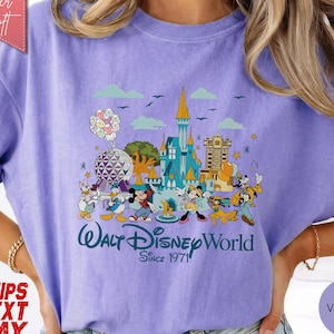 K&ouml;nnte beinhalten: Ein violettes T-Shirt mit einer farbenfrohen Grafik von Disney-Figuren und Wahrzeichen. Das Design umfasst Micky Maus, Donald Duck und ein Schloss mit dem Text "Walt Disney World Since 1971". Das Shirt hat kurze &Auml;rmel und eine lockere Passform.