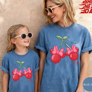 May include: Two people wearing matching blue jean t-shirts. Each shirt features a cartoon cherry design with pink bows and the Disney logo. The shirts are a relaxed fit and have rolled-up sleeves.