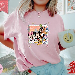 Może przedstawiać: Jasnor&oacute;żowa koszulka z kolorową grafiką postaci Disneya: Myszki Miki, Myszki Minnie, Goofy'ego, Kaczora Donalda i Daisy Duck. Koszulka ma okrągły dekolt i kr&oacute;tkie rękawy. Napis "Super Soft" znajduje się na r&oacute;żowym banerze.