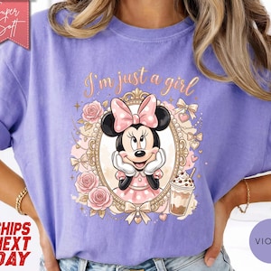 Może przedstawiać: Fioletowy t-shirt z grafiką Myszki Minnie w ozdobnej złotej owalnej ramce. Nad ramką znajduje się napis "I'm just a girl". Wz&oacute;r zawiera r&oacute;żowe r&oacute;że, kokardki i kubek z napojem. Czerwona metka w rogu zawiera napis "Super Soft".