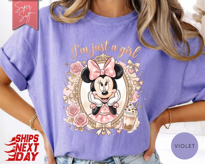 Może przedstawiać: Fioletowy t-shirt z grafiką Myszki Minnie w ozdobnej złotej owalnej ramce. Nad ramką znajduje się napis "I'm just a girl". Wz&oacute;r zawiera r&oacute;żowe r&oacute;że, kokardki i kubek z napojem. Czerwona metka w rogu zawiera napis "Super Soft".