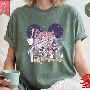 May include: Moss green t-shirt featuring a graphic of Disney characters and the word "Epcot" in pink and white. The design includes Mickey Mouse, Donald Duck, and Goofy. The shirt has rolled-up sleeves and a relaxed fit.