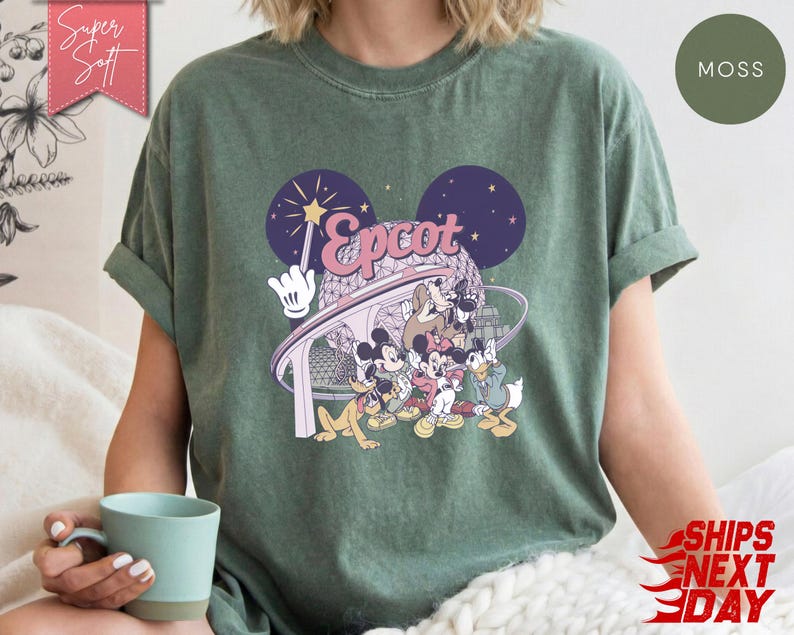 May include: Moss green t-shirt featuring a graphic of Disney characters and the word "Epcot" in pink and white. The design includes Mickey Mouse, Donald Duck, and Goofy. The shirt has rolled-up sleeves and a relaxed fit.