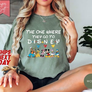 K&ouml;nnte beinhalten: Ein olivgr&uuml;nes T-Shirt mit dem Text "The One Where They Go To Disney" und Disney-Figuren. Das Shirt hat eine lockere Passform und kurze &Auml;rmel. Das Bild enth&auml;lt auch den Text "Super Soft" und "Ships Next Day".