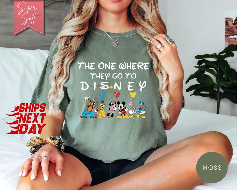 K&ouml;nnte beinhalten: Ein olivgr&uuml;nes T-Shirt mit dem Text "The One Where They Go To Disney" und Disney-Figuren. Das Shirt hat eine lockere Passform und kurze &Auml;rmel. Das Bild enth&auml;lt auch den Text "Super Soft" und "Ships Next Day".