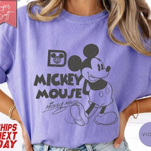 May include: A violet-colored t-shirt featuring a distressed graphic of Mickey Mouse. The design includes the words "Mickey Mouse" in a bold font, with Mickey's image to the right. The shirt has rolled-up sleeves.