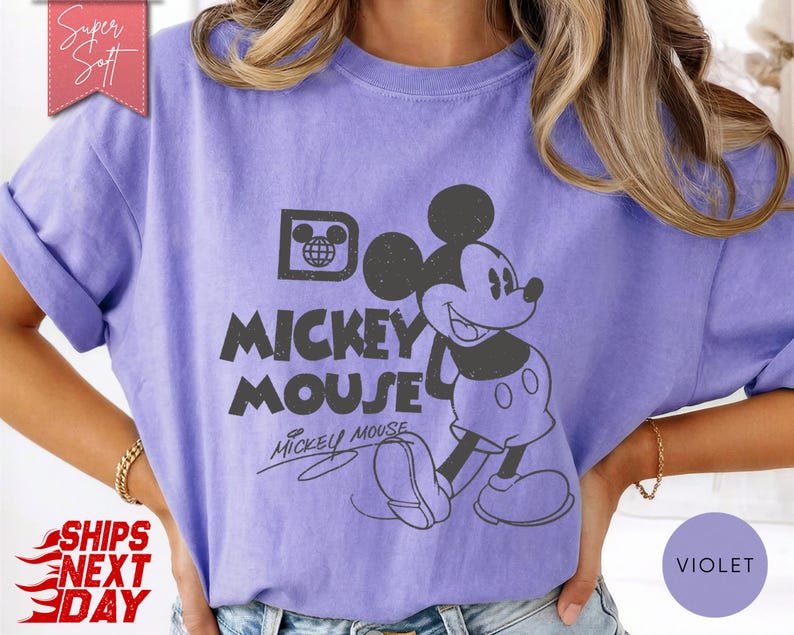 May include: A violet-colored t-shirt featuring a distressed graphic of Mickey Mouse. The design includes the words "Mickey Mouse" in a bold font, with Mickey's image to the right. The shirt has rolled-up sleeves.