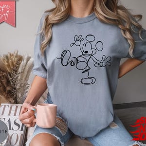 May include: A light blue t-shirt with a black line drawing of Mickey Mouse. The shirt has the words "Love" and "Ours" in a script font. The words "Super Soft" are in a pink banner.