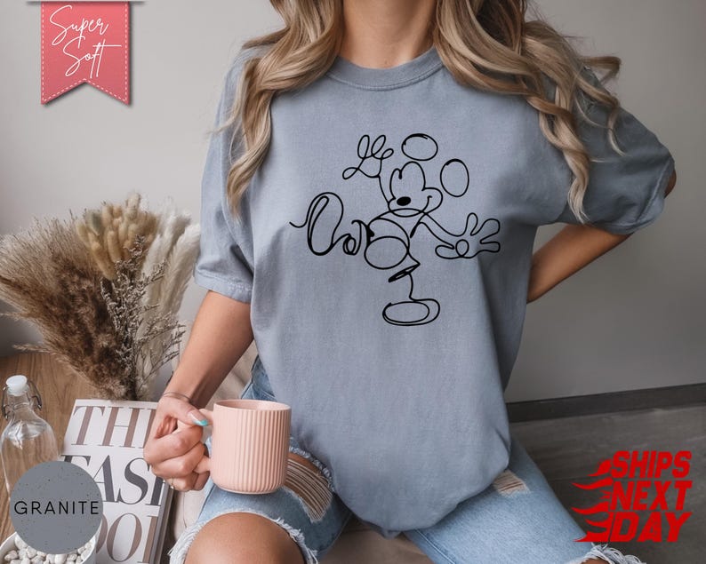 May include: A light blue t-shirt with a black line drawing of Mickey Mouse. The shirt has the words "Love" and "Ours" in a script font. The words "Super Soft" are in a pink banner.
