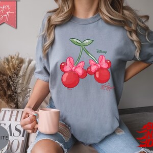 May include: A light blue t-shirt featuring a cartoon cherry design with Minnie Mouse bows. The cherries are red with pink bows and green stems. The shirt has the text "Disney Minnie Mouse" printed on it. The shirt is labeled "Super Soft".