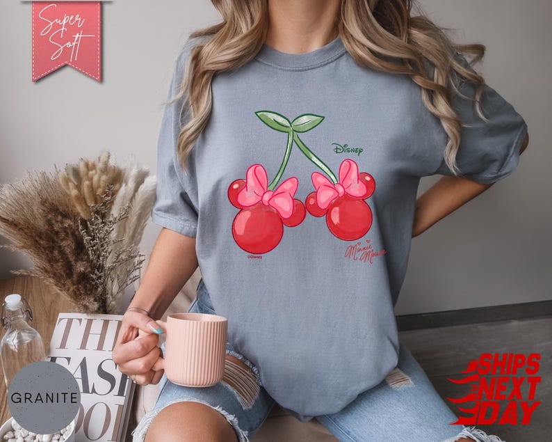 May include: A light blue t-shirt featuring a cartoon cherry design with Minnie Mouse bows. The cherries are red with pink bows and green stems. The shirt has the text "Disney Minnie Mouse" printed on it. The shirt is labeled "Super Soft".