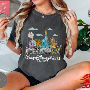 K&ouml;nnte beinhalten: Dunkelgraues T-Shirt mit einer farbenfrohen Grafik von Walt Disney World-Charakteren und Wahrzeichen, darunter Cinderella's Schloss und Spaceship Earth. Der Text "Walt Disney World Since 1971" ist darunter gedruckt. Das Shirt hat die Farbe Pfeffer.