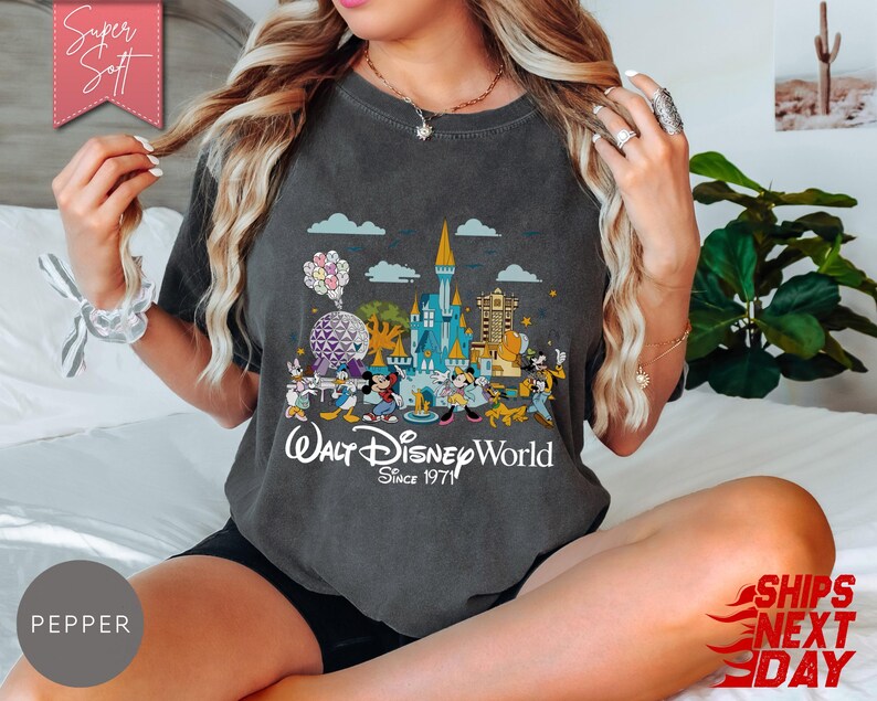 K&ouml;nnte beinhalten: Dunkelgraues T-Shirt mit einer farbenfrohen Grafik von Walt Disney World-Charakteren und Wahrzeichen, darunter Cinderella's Schloss und Spaceship Earth. Der Text "Walt Disney World Since 1971" ist darunter gedruckt. Das Shirt hat die Farbe Pfeffer.