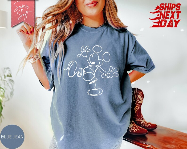 May include: Blue jean-colored t-shirt featuring a white line drawing of Mickey Mouse. The shirt has short sleeves and a crew neck. The words "Super Soft" are on a pink banner in the upper left corner. The words "Ships Next Day" are in red on the upper right.