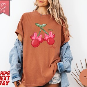 May include: A rust-colored t-shirt featuring a Minnie Mouse cherry design. The graphic includes two red cherries with pink bows and green leaves, with the Disney and Minnie Mouse logos. The shirt has rolled-up sleeves.