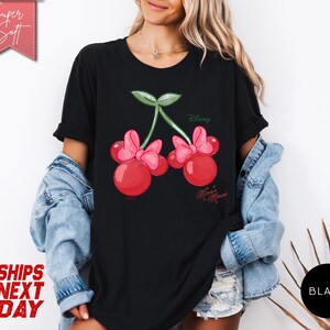 May include: Black t-shirt featuring a Minnie Mouse cherry design. The graphic includes two red cherries with pink bows, green leaves, and the Disney and Minnie Mouse logos. The shirt has short sleeves.