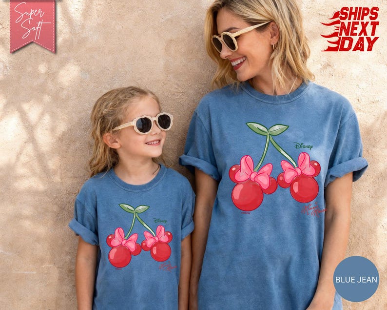 May include: Two people wearing matching blue jean t-shirts. Each shirt features a cartoon cherry design with pink bows and the Disney logo. The shirts are a relaxed fit and have rolled-up sleeves.