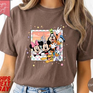 Może przedstawiać: Koszulka w kolorze espresso z kolorową grafiką Myszki Miki, Myszki Minnie, Goofy'ego, Kaczora Donalda, Daisy Duck i Pluto. Grafika jest obramowana pastelową obw&oacute;dką i gwiazdkami. Koszulka ma napis "Super Soft" u g&oacute;ry po lewej.