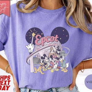 May include: Violet-colored t-shirt featuring a graphic of Disney characters and the word "Epcot." The design includes Mickey Mouse, Donald Duck, and other characters. The shirt has rolled-up sleeves and a relaxed fit.