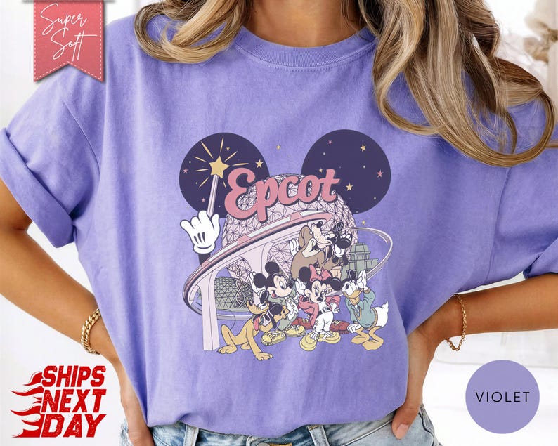 May include: Violet-colored t-shirt featuring a graphic of Disney characters and the word "Epcot." The design includes Mickey Mouse, Donald Duck, and other characters. The shirt has rolled-up sleeves and a relaxed fit.