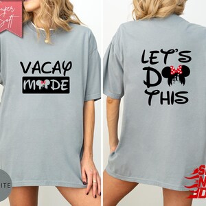 May include: A light gray t-shirt with the words "VACAY MODE" printed on the front and "LET'S DO THIS" on the back. The front design includes a black rectangle with a Mickey Mouse head silhouette. The back design features a Mickey Mouse head with a red bow.