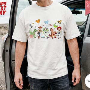 May include: A white short-sleeved t-shirt featuring a graphic print of various animated toy characters including a cowboy, a space ranger, a cowgirl, a dinosaur, a pig, and a horse, each holding a colourful Mickey Mouse-shaped balloon.