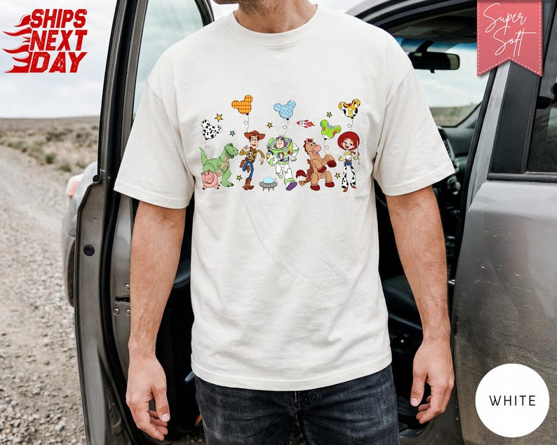 May include: A white short-sleeved t-shirt featuring a graphic print of various animated toy characters including a cowboy, a space ranger, a cowgirl, a dinosaur, a pig, and a horse, each holding a colourful Mickey Mouse-shaped balloon.