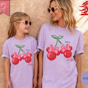 May include: Two light purple t-shirts featuring a Minnie Mouse cherry design. The shirts have rolled-up sleeves and are worn by a mother and daughter. The design includes red cherries, pink bows, and green leaves. The word "Disney" is printed on the shirt.