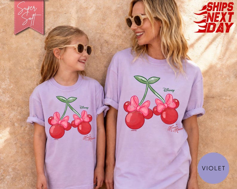 May include: Two light purple t-shirts featuring a Minnie Mouse cherry design. The shirts have rolled-up sleeves and are worn by a mother and daughter. The design includes red cherries, pink bows, and green leaves. The word "Disney" is printed on the shirt.