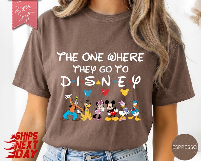 K&ouml;nnte beinhalten: Braunes T-Shirt mit dem wei&szlig;en Text "The One Where They Go To Disney". Das Design zeigt Zeichentrickfiguren wie Micky und Minnie Maus, Donald und Daisy Duck sowie Goofy. Bunte Luftballons sind ebenfalls abgebildet.