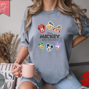 May include: A light blue t-shirt featuring the text "Disney Mickey and Friends" with colorful character portraits. The shirt has a relaxed fit and short sleeves. The shirt is made of a soft material.