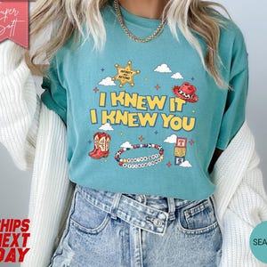 May include: A seafoam green short-sleeve t-shirt featuring a graphic print with the text I KNEW IT I KNEW YOU, a yellow sheriff star, a red cowboy hat, a red cowboy boot, a beaded bracelet, and alphabet blocks against a background of clouds.