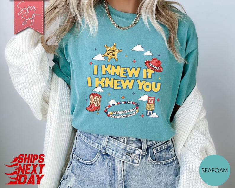 May include: A seafoam green short-sleeve t-shirt featuring a graphic print with the text I KNEW IT I KNEW YOU, a yellow sheriff star, a red cowboy hat, a red cowboy boot, a beaded bracelet, and alphabet blocks against a background of clouds.