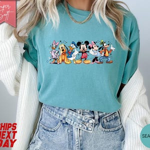 Puede incluir: Camiseta verde agua con un dise&ntilde;o de personajes de Disney: Mickey, Minnie, Donald, Daisy, Goofy y Pluto. La camiseta tiene cuello redondo y mangas cortas. Las palabras "Super Soft" est&aacute;n en una pancarta rosa. Hecha de material suave.